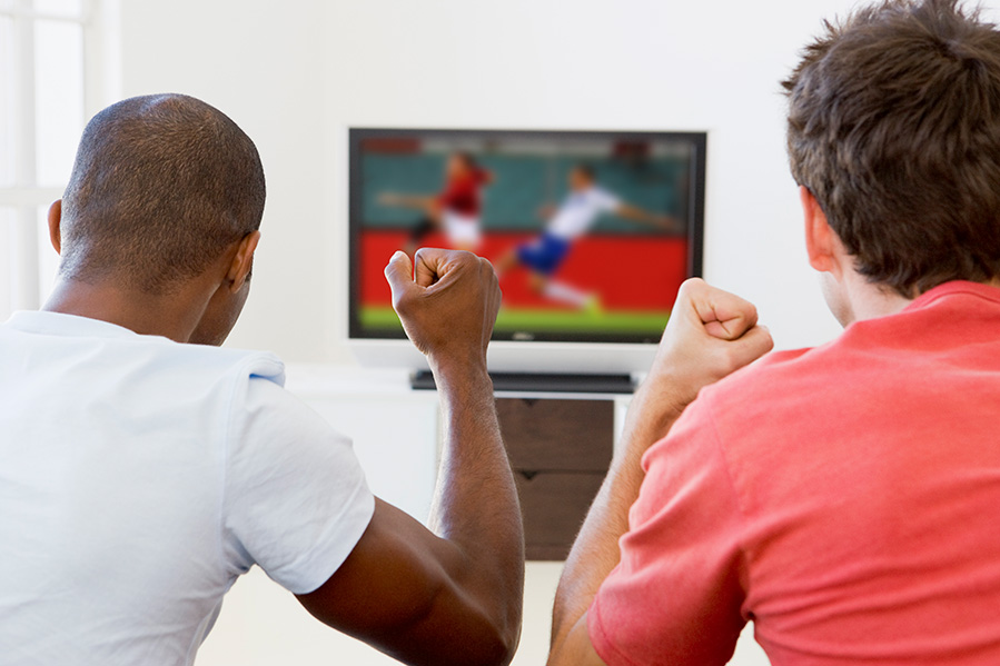 View from behind of two friends sitting on a couch and watching a soccer game both excited.